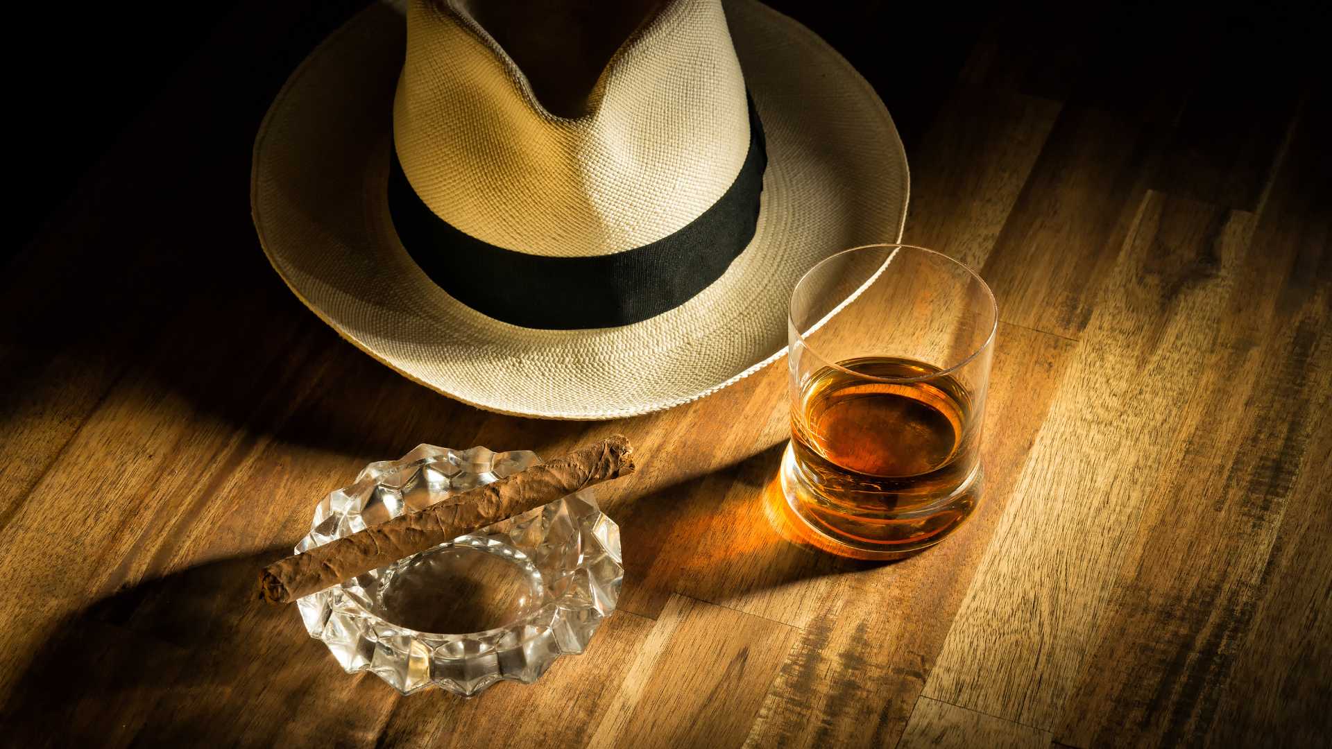 Un bicchiere di rum, un sigaro e un cappello di Panama su un tavolo in legno, evocando un’atmosfera elegante e sofisticata, ideale per gustare un distillato pregiato.